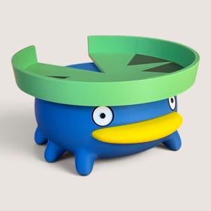 Puede incluir: Un recipiente azul con forma de criatura, pico amarillo y dos ojos blancos. La parte superior tiene una tapa de hoja de nenúfar verde con un corte triangular. La base tiene cuatro pequeñas patas azules y un acabado liso y mate.