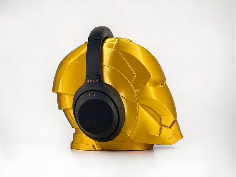 K&ouml;nnte beinhalten: Schwarze Over-Ear-Kopfh&ouml;rer mit dem Schriftzug "SONY" an der Seite, die auf einem goldfarbenen, helmf&ouml;rmigen St&auml;nder ruhen.