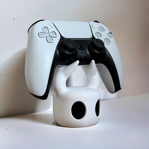 K&ouml;nnte beinhalten: Ein wei&szlig;er PlayStation 5 DualSense-Controller steht auf einem wei&szlig;en 3D-gedruckten St&auml;nder, der wie der Ritter aus dem Videospiel Hollow Knight geformt ist.