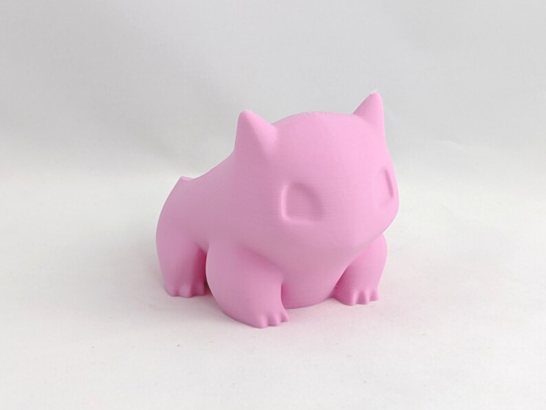May include: A pink 3D printed figurine of a Pokemon character. The figurine is sitting on a white surface.