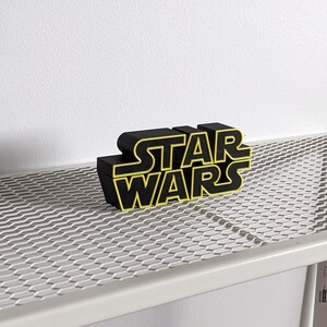 May include: Black "Star Wars" logo with yellow outlines, sitting on a white metal shelf with a wire mesh surface.