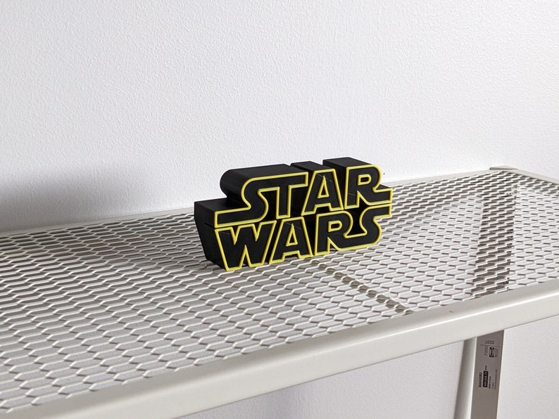 May include: Black "Star Wars" logo with yellow outlines, sitting on a white metal shelf with a wire mesh surface.