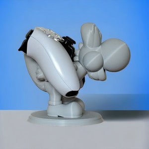 May include: A white 3D printed figurine of a gray Yoshi holding a white video game controller. The Yoshi is standing on a round base.