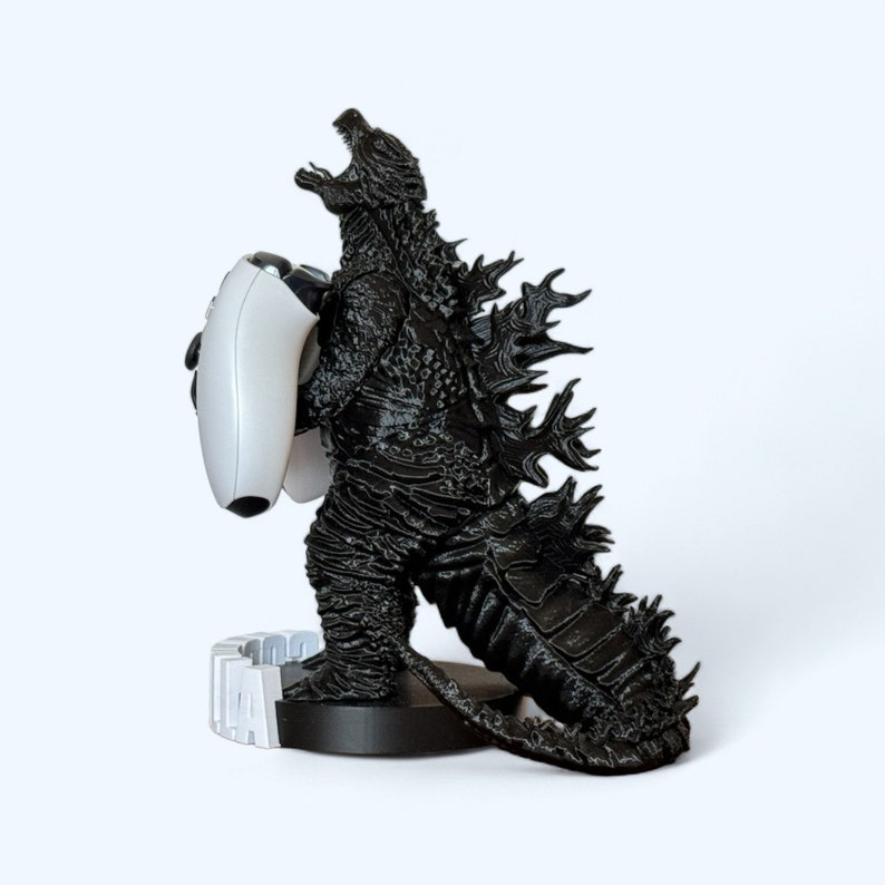 May include: A black Godzilla figurine holding a white video game controller. The figurine is standing on a black base with a white base.