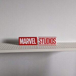 K&ouml;nnte beinhalten: Zwei rote Bl&ouml;cke mit wei&szlig;en Buchstaben. Der linke Block sagt "MARVEL" und der rechte Block sagt "STUDIOS".