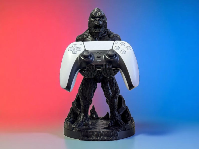 May include: Black gorilla-shaped controller stand holding a white and black game controller. The stand is on a round base with a textured surface. The background gradient transitions from red to blue.