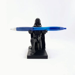 May include: A black Darth Vader pen holder with a blue pen. The pen holder is a detailed figurine of Darth Vader kneeling, holding the pen horizontally. The pen is blue with a silver tip.
