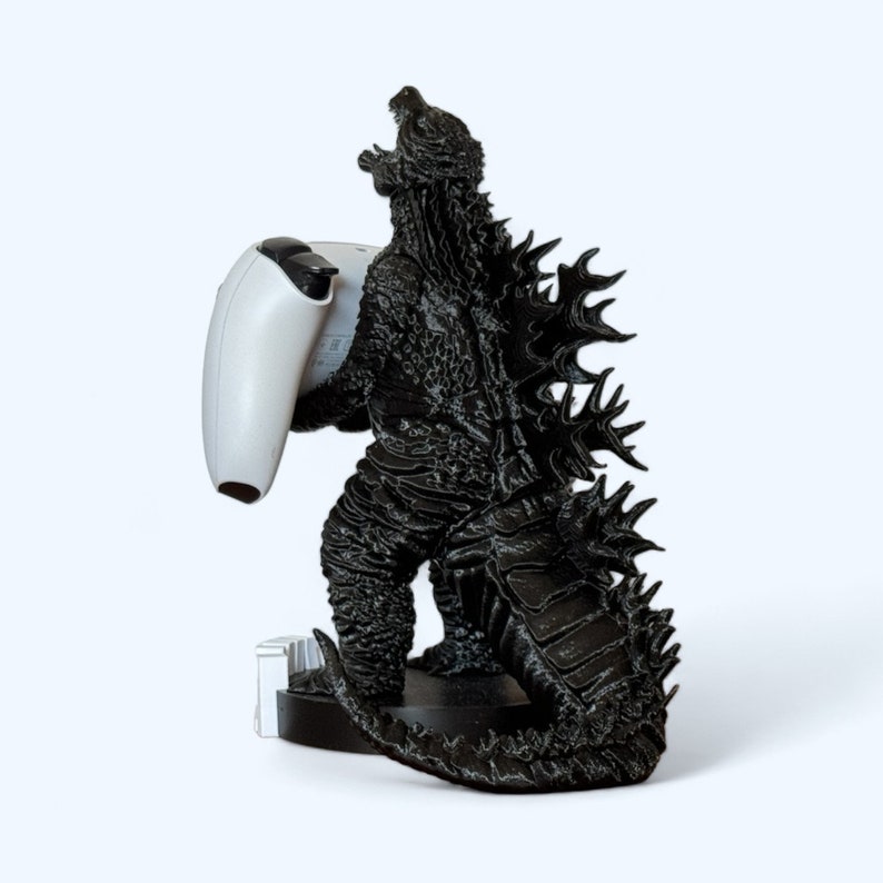 May include: A black Godzilla figurine holding a white video game controller. The figurine is standing on a black base.