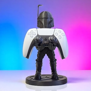 May include: A black and white PlayStation 5 controller holder featuring a Mandalorian helmet design. The holder is black with a round base and holds a white and black controller. The background has blue, pink, and purple hues.