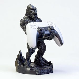 May include: A black gorilla statue holding a white and black video game controller. The statue has a textured finish and is mounted on a black base. The controller has a sleek, modern design.