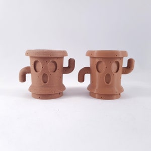 May include: Two brown ceramic planters shaped like cacti with a face and arms. The planters are 3D printed and have a textured surface.