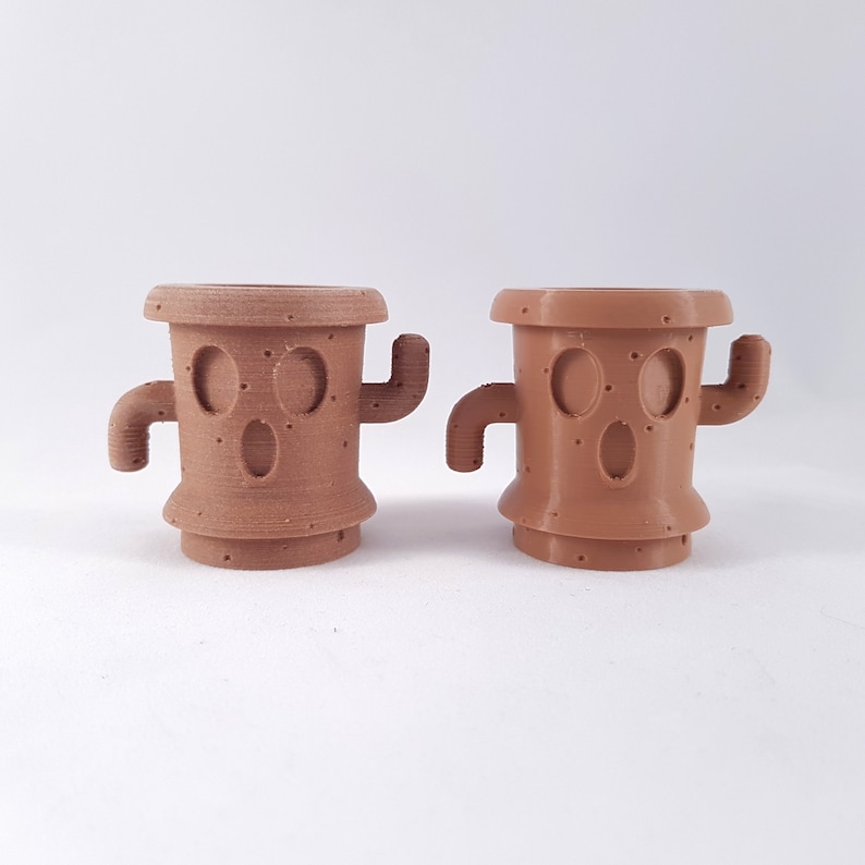 May include: Two brown ceramic planters shaped like cacti with a face and arms. The planters are 3D printed and have a textured surface.