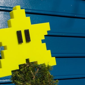 Puede incluir: Un objeto amarillo brillante en forma de estrella con dos rectángulos negros para los ojos, sentado en un arbusto verde sobre un fondo azul.