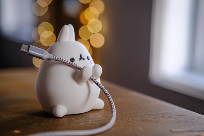 May include: White ceramic bunny figurine holding a gray braided USB cable with a USB-C connector. The bunny has black eyes and a pink nose.