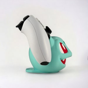 May include: A teal green and red 3D printed stand for a video game controller, shaped like a cartoon character. The stand is designed to hold a white video game controller.