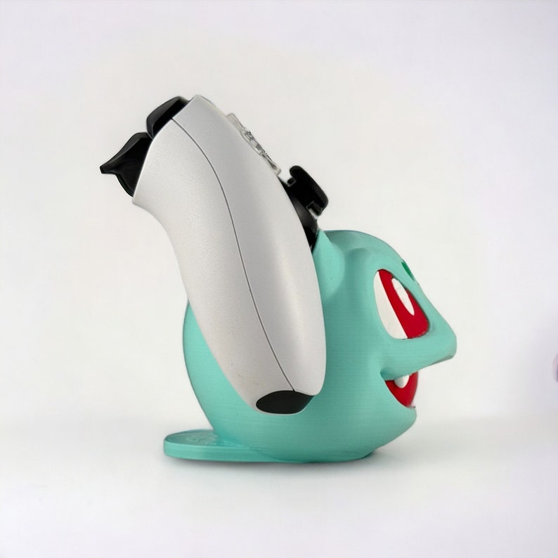 May include: A teal green and red 3D printed stand for a video game controller, shaped like a cartoon character. The stand is designed to hold a white video game controller.