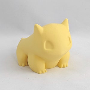 May include: A yellow 3D printed figurine of a Pokemon character, resembling a small, round creature with large ears and a wide mouth.
