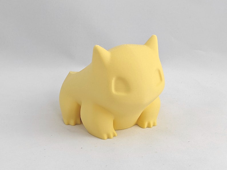May include: A yellow 3D printed figurine of a Pokemon character, resembling a small, round creature with large ears and a wide mouth.