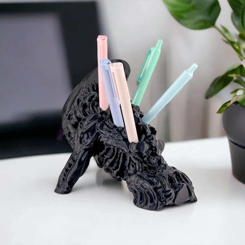 May include: A black, 3D printed, skull-shaped desk organizer with a textured surface holds five pens. The pens are pink, light blue, and a darker shade of blue.