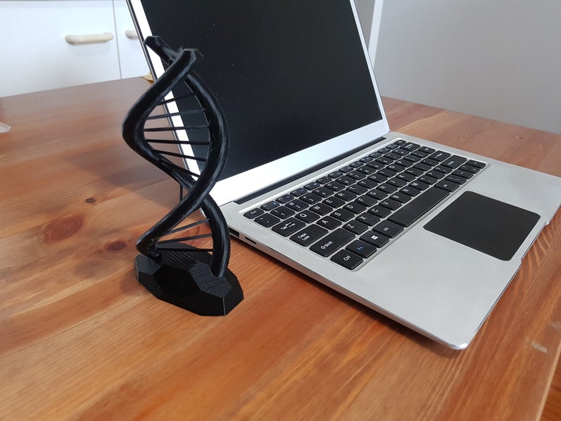 Op de afbeelding: Een zwart 3D-geprint model van een DNA-dubbele helix op een houten oppervlak naast een zilveren laptop.