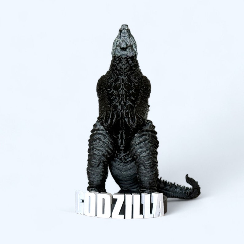 May include: A black Godzilla figurine with a textured surface, sitting on a white base with the word "GODZILLA" printed in white.