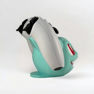May include: A white and teal 3D printed game controller stand shaped like a Pokemon character. The stand has a red eye and a toothy grin.