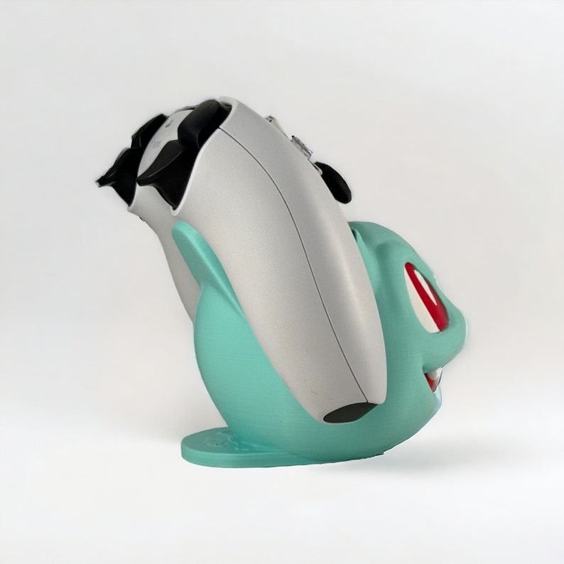 May include: A white and teal 3D printed game controller stand shaped like a Pokemon character. The stand has a red eye and a toothy grin.