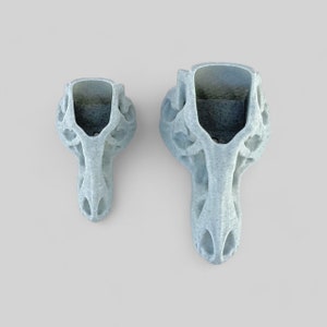 Op de afbeelding: Twee witte keramische dinosaurus schedel potten, een groter dan de andere. De potten zijn gedetailleerd met een getextureerd, bot-achtig oppervlak.