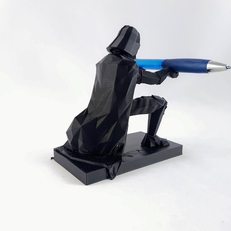 May include: A black 3D-printed pen holder shaped like Darth Vader, kneeling and holding a blue pen. The pen holder is on a rectangular base, and the figure has a geometric design.