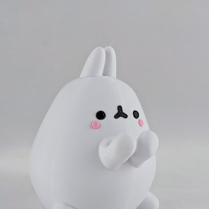 May include: White bunny figurine with black eyes and pink cheeks. The bunny is holding its paws together in front of its chest.