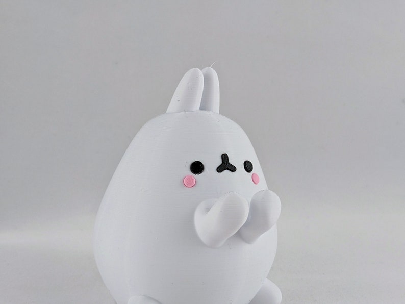 May include: White bunny figurine with black eyes and pink cheeks. The bunny is holding its paws together in front of its chest.