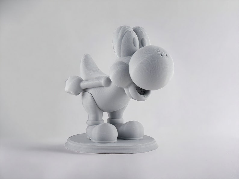 May include: A white 3D printed figurine of a cartoon dinosaur character with a large head and a wide smile. The figurine is standing on a circular base.
