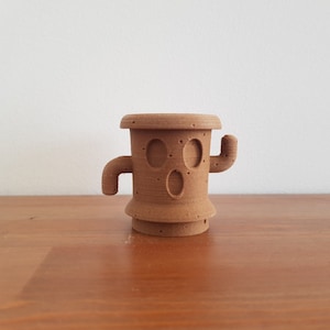 May include: A brown 3D printed cactus figurine with a face and arms. The cactus is standing on a wooden surface.