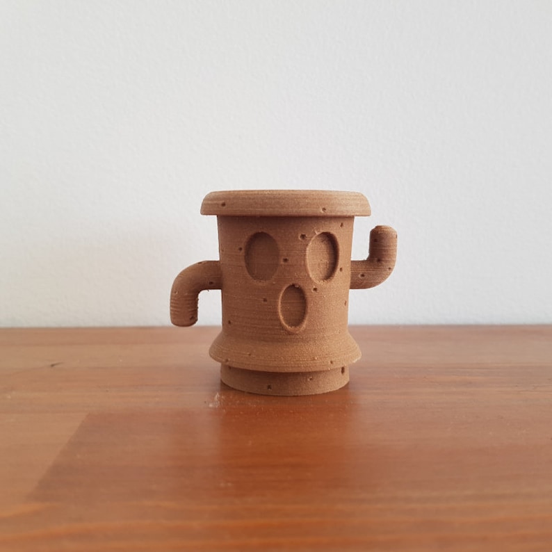 May include: A brown 3D printed cactus figurine with a face and arms. The cactus is standing on a wooden surface.