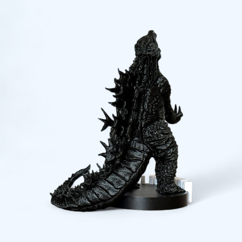 May include: Black and grey detailed Godzilla figurine with a spiky back and tail. The figurine is standing on a black base.
