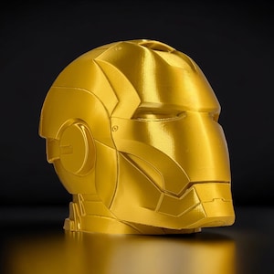 K&ouml;nnte beinhalten: Eine goldfarbene, 3D-gedruckte Nachbildung eines Superhelden-Helms. Der Helm hat ein elegantes, futuristisches Design mit filigranen Details und einer glatten, metallischen Oberfl&auml;che.