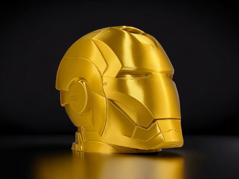 K&ouml;nnte beinhalten: Eine goldfarbene, 3D-gedruckte Nachbildung eines Superhelden-Helms. Der Helm hat ein elegantes, futuristisches Design mit filigranen Details und einer glatten, metallischen Oberfl&auml;che.