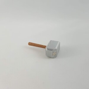 May include: A small, silver-colored hammer with a brown wooden handle. The hammer is a replica of Thor's hammer, Mjolnir, from the Marvel comics and movies.