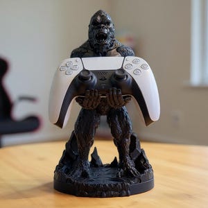 May include: A black and gray gorilla-shaped controller stand holding a white and black game controller. The stand is on a wooden table. The controller has a black center and white sides.