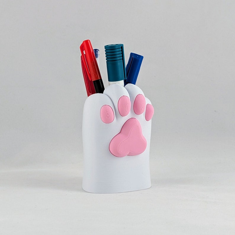 Puede incluir: Porta-bol&iacute;grafos de cer&aacute;mica blanca en forma de pata con huellas de patas rosas. El porta-bol&iacute;grafos contiene cuatro bol&iacute;grafos y marcadores, incluyendo un bol&iacute;grafo rojo, un bol&iacute;grafo azul, un marcador negro y un marcador azul.