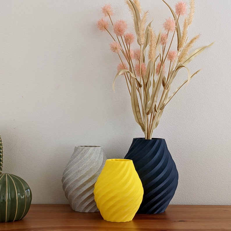 May include: Three ceramic vases in different colors and designs. A gray vase with a textured surface, a yellow vase with a swirl pattern, and a dark blue vase with a swirl pattern. The vases are arranged on a wooden surface.