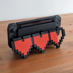 May include: A black Nintendo Switch console with a 3D printed stand shaped like a pixelated heart. The stand is red and black.