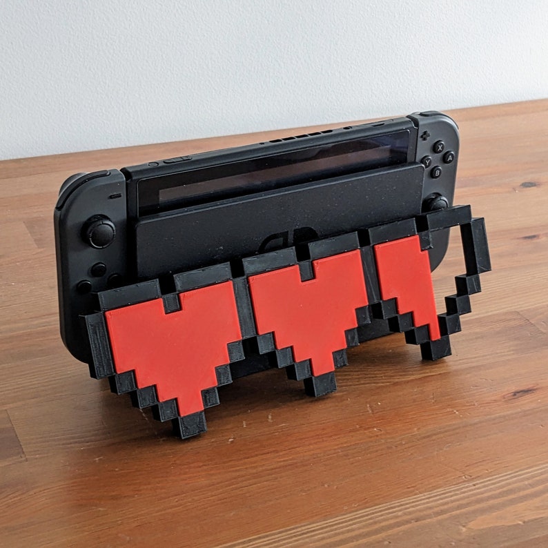 May include: A black Nintendo Switch console with a 3D printed stand shaped like a pixelated heart. The stand is red and black.