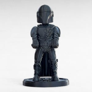 Pode incluir: Uma estatueta cinza escuro de um personagem blindado, com capacete e capa. A figura est&aacute; em p&eacute; sobre uma base circular. A armadura tem texturas detalhadas e a figura est&aacute; em pose com os punhos cerrados.