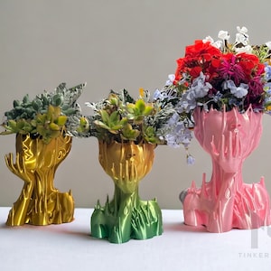 May include: Three 3D printed planters shaped like human heads, each with a different colour. The planter on the left is gold, the middle planter is green, and the planter on the right is pink. The planters are filled with plants.