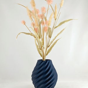 K&ouml;nnte beinhalten: Eine dunkelblaue Vase mit einem wirbelnden, strukturierten Design h&auml;lt einen Strau&szlig; getrockneter Pampasgr&auml;ser mit rosa Bl&uuml;tenk&ouml;pfen.