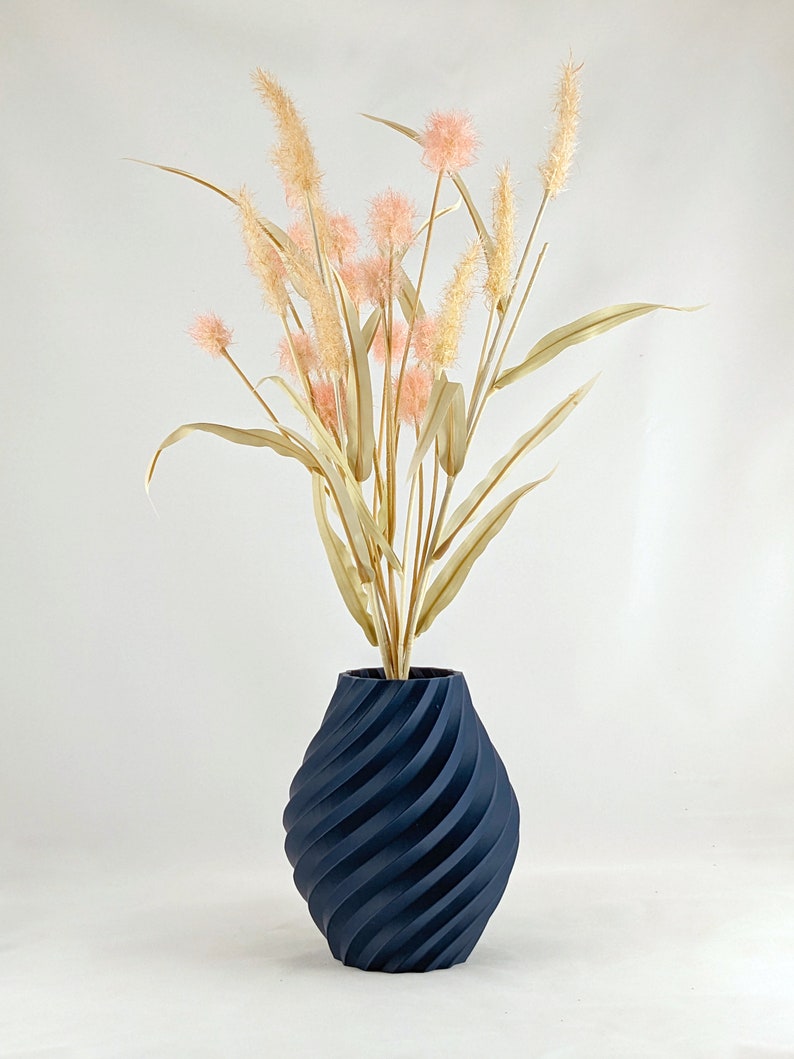K&ouml;nnte beinhalten: Eine dunkelblaue Vase mit einem wirbelnden, strukturierten Design h&auml;lt einen Strau&szlig; getrockneter Pampasgr&auml;ser mit rosa Bl&uuml;tenk&ouml;pfen.