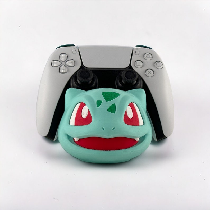 May include: A white and black video game controller with a green and blue Bulbasaur character attached to the front.