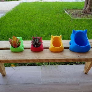 May include: Four colorful ceramic planters in the shape of a cartoon animal, sitting on a wooden bench. The planters are green, red, yellow, and blue.