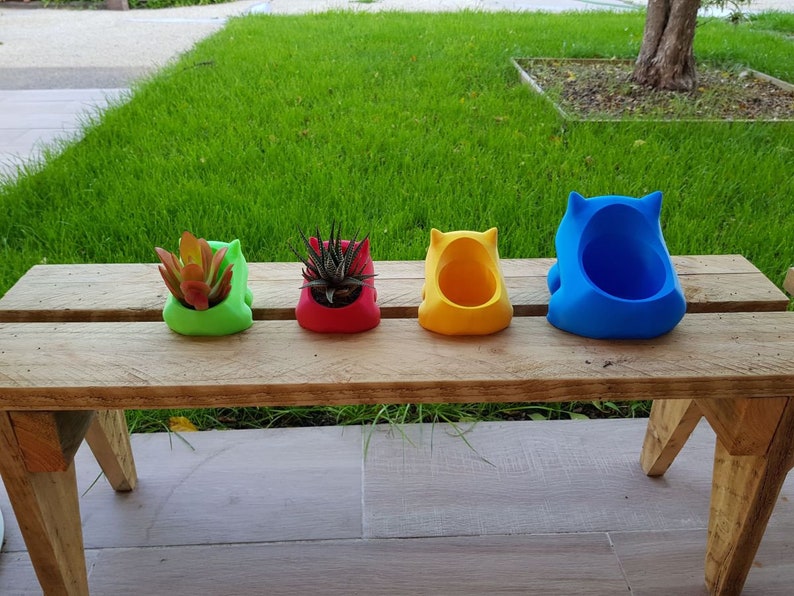 May include: Four colorful ceramic planters in the shape of a cartoon animal, sitting on a wooden bench. The planters are green, red, yellow, and blue.